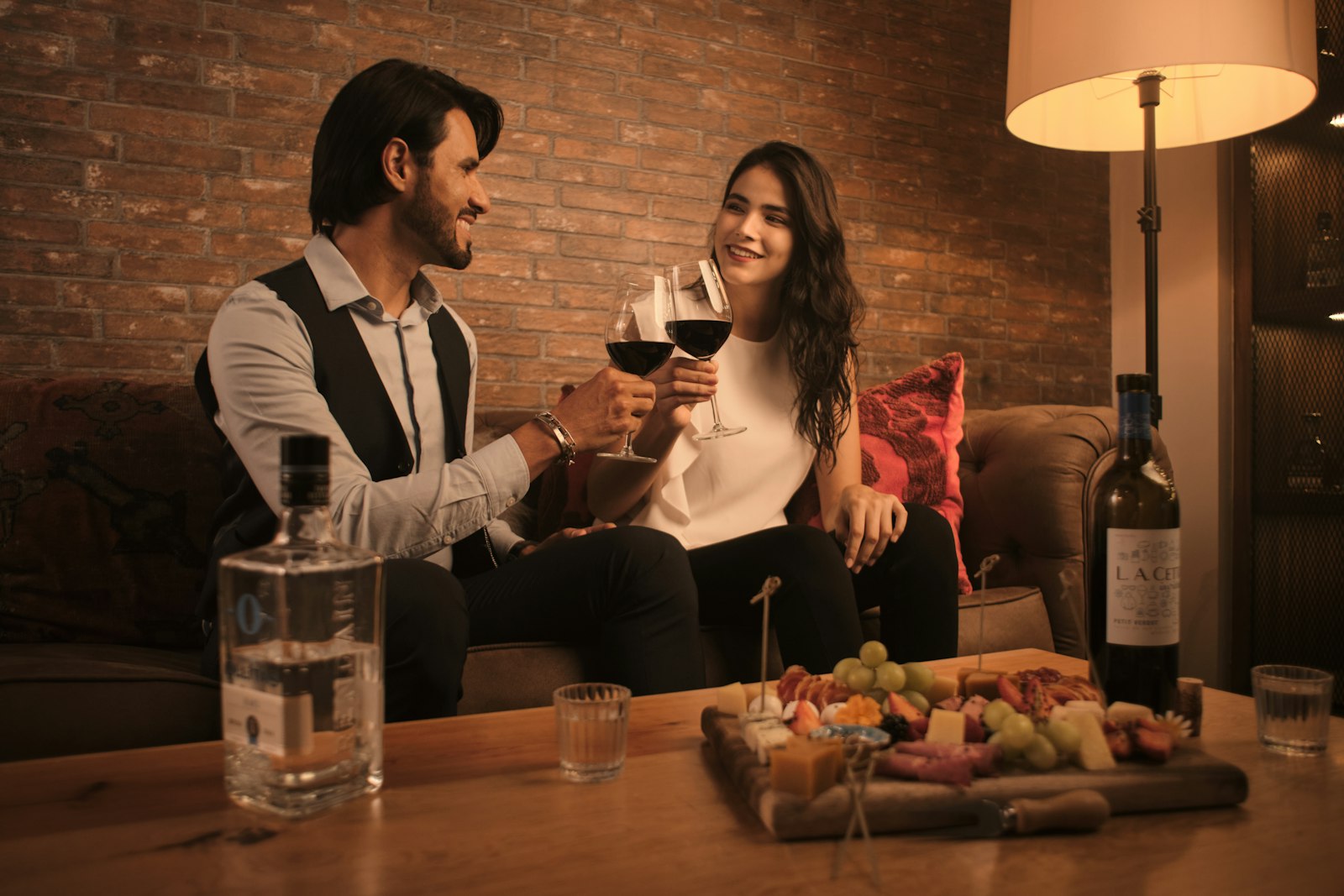 Casal brindando com taças de vinho tinto na sala de casa, com luz baixa